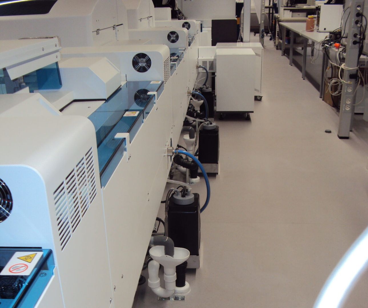 Eine Reihe weißer Laborgeräte mit blauen Akzenten, mit Ventilatoren und angeschlossenen Rohren, in einer sauberen Laborumgebung.