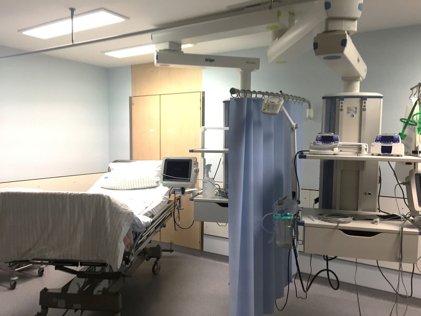 Ein Patientenzimmer mit einem Krankenhausbett, medizinischen Geräten und blauen Vorhängen in einer hellen, sauberen Umgebung.
