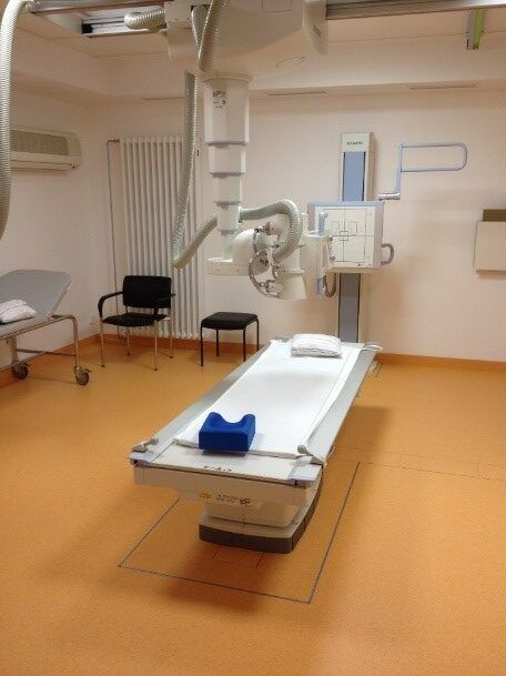 Ein sauberer, gut beleuchteter medizinischer Untersuchungsraum mit einem Tisch mit Bettwäsche, einem blauen Stützkissen und einem Radiologiegerät über dem Kopf.